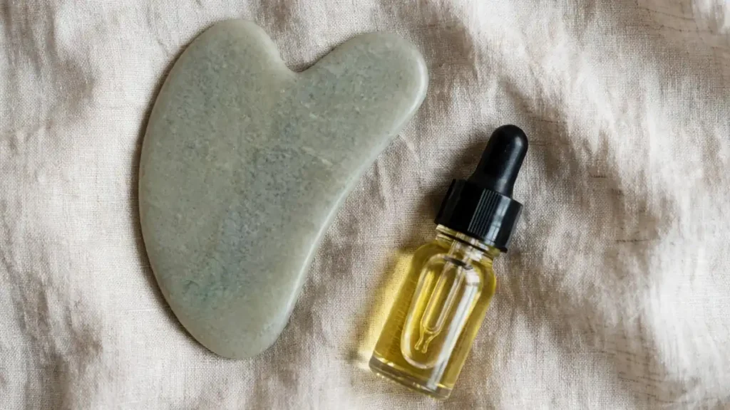 Smooth pale green stone gua sha tool beside a small unlabelled dropper bottle of facial oil on soft beige linen