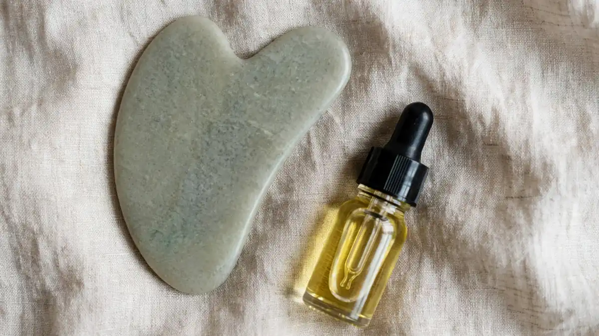 Smooth pale green stone gua sha tool beside a small unlabelled dropper bottle of facial oil on soft beige linen